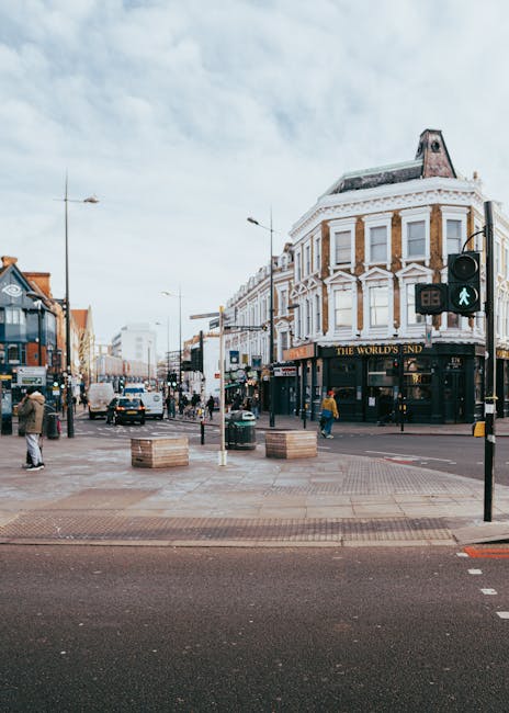 A busy street scene in Camden Town, London, showing a corner with historic, multi-story buildings featuring white facades and large windows. Pedestrians are walking on the pavement; some are crossing 
