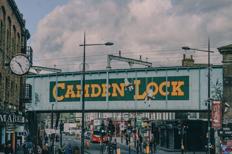 A busy street scene in Camden Town, London, showing a corner with historic, multi-story buildings featuring white facades and large windows. Pedestrians are walking on the pavement; some are crossing 